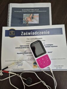 Zaświadczenie o ukończeniu kursu logopedycznego z elektrostymulacji leżące na biurku, obok bindowanego podręcznika i przenośnego elektrostymulatora Logotens z podłączonymi przewodami.