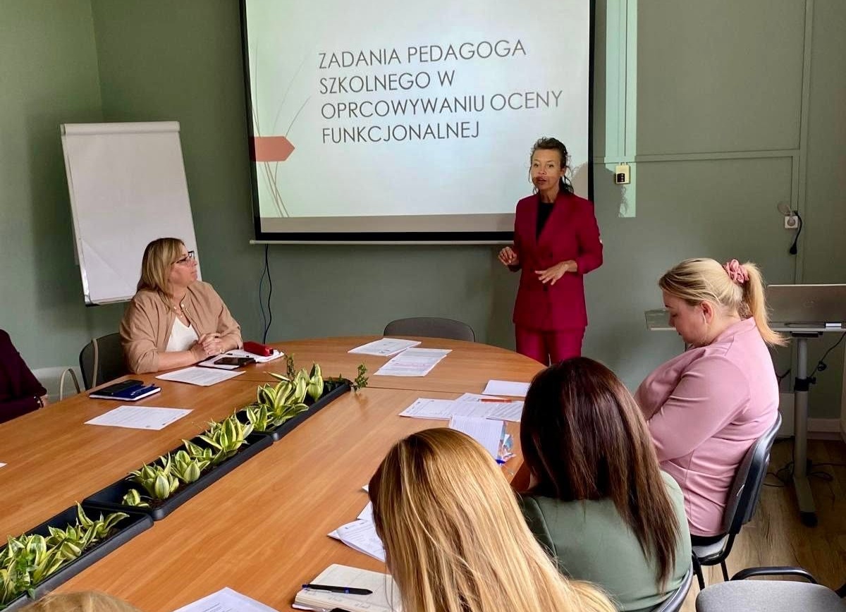 Kobieta w bordowym garniturze prowadzi prezentację dla grupy osób siedzących przy dużym stole konferencyjnym. Na ekranie za nią widnieje napis: „ZADANIA PEDAGOGA SZKOLNEGO W OPRACOWYWANIU OCENY FUNKCJONALNEJ”.